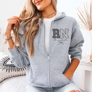 May include: A heather gray zip-up hoodie featuring a white undershirt. The left chest displays the letters RN in a block font with a gradient effect, accompanied by the name Elizabeth in cursive script and a small bow graphic underneath.