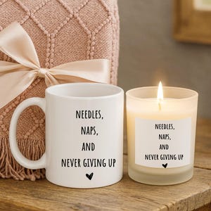 Puede incluir: Una taza de cerámica blanca y una vela encendida con el texto "NEEDLES, NAPS, AND NEVER GIVING UP" se exhiben sobre una superficie de madera. Una manta rosa con un lazo está en el fondo. La vela está en un recipiente de vidrio esmerilado.