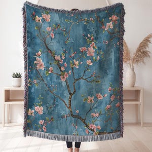 May include: A rectangular woven throw blanket featuring a deep blue background with a pattern of brown branches and light pink blossoms. The blanket has a textured fringe along all four edges, creating a decorative border around the floral design.