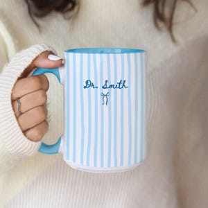 May include: A white ceramic mug featuring vertical light blue stripes, a solid light blue handle, and a light blue interior. The text Dr. Smith is printed in dark blue cursive script above a small dark blue bow icon on the front of the mug.