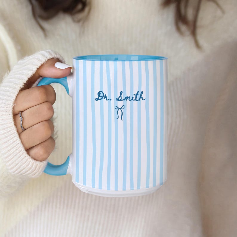May include: A white ceramic mug featuring vertical light blue stripes, a solid light blue handle, and a light blue interior. The text Dr. Smith is printed in dark blue cursive script above a small dark blue bow icon on the front of the mug.