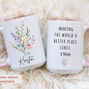 May include: Two white ceramic mugs with pink handles and rims. One mug features a floral design and the name "Kristin." The other mug has the text "MAKING THE WORLD A BETTER PLACE SINCE 1966."