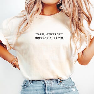 May include: A cream-coloured short-sleeve t-shirt featuring the black text HOPE, STRENGTH SCIENCE & FAITH printed in a typewriter-style font across the centre of the chest. The garment has a crew neckline and a relaxed fit.
