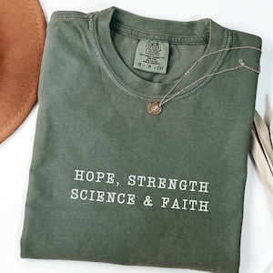 May include: An olive green crew neck t-shirt featuring the white text "HOPE, STRENGTH SCIENCE & FAITH" printed on the chest. The shirt has a Comfort Colors brand label at the inner neckline indicating size S, 100% cotton, and made in Honduras.