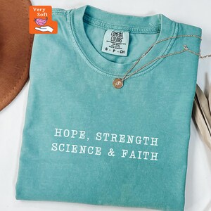 May include: A teal short-sleeved t-shirt with a crew neckline featuring the white printed text HOPE, STRENGTH SCIENCE & FAITH. The shirt has a Comfort Colors brand label at the inner neck and is displayed with a delicate gold-tone necklace.