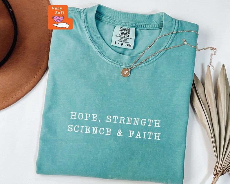 May include: A teal short-sleeved t-shirt with a crew neckline featuring the white printed text HOPE, STRENGTH SCIENCE & FAITH. The shirt has a Comfort Colors brand label at the inner neck and is displayed with a delicate gold-tone necklace.