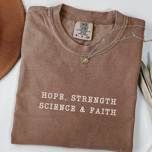 May include: A brown short-sleeved t-shirt with a crew neckline and a Comfort Colors brand label. The shirt features the white text HOPE, STRENGTH SCIENCE & FAITH printed on the front. A silver-toned necklace with a small circular pendant rests on the fabric.