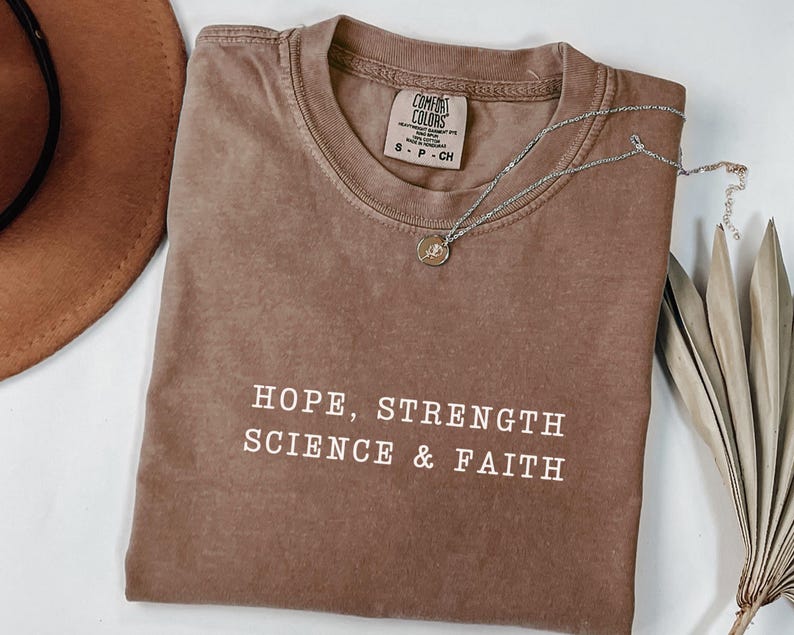 May include: A brown short-sleeved t-shirt with a crew neckline and a Comfort Colors brand label. The shirt features the white text HOPE, STRENGTH SCIENCE & FAITH printed on the front. A silver-toned necklace with a small circular pendant rests on the fabric.