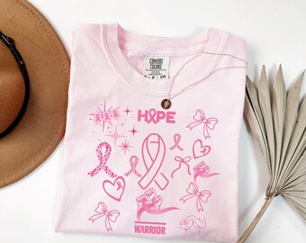 May include: A light pink t-shirt with the word "HOPE" and various pink ribbon graphics. The shirt also features the word "WARRIOR" and a necklace with a pendant. The shirt is folded on a white surface.