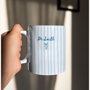 May include: A white ceramic mug featuring vertical light blue stripes. The text Dr. Smith is printed in dark blue script font above a small dark blue bow icon. The mug has a white handle and a smooth, cylindrical body design.