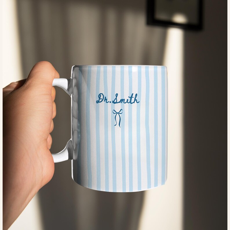 May include: A white ceramic mug featuring vertical light blue stripes. The text Dr. Smith is printed in dark blue script font above a small dark blue bow icon. The mug has a white handle and a smooth, cylindrical body design.