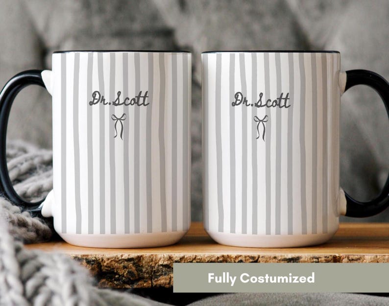 May include: Two white ceramic mugs featuring a vertical gray striped pattern. Each mug displays the text Dr. Scott in a dark gray script font, positioned above a small, simple line-drawn bow icon. The mugs have solid black handles and rims.