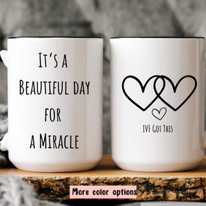 May include: Two white ceramic mugs with black rims. One mug reads "It's a beautiful day for a miracle." The other mug has two intertwined hearts and the text "IVF got this."