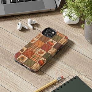 May include: A smartphone case featuring a patchwork design of autumn-themed squares. The pattern includes quilted textures, orange pumpkins, maple leaves, sunflowers, and plaid prints in shades of orange, brown, olive green, and tan colours.