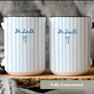 May include: Two white ceramic mugs with a light blue vertical striped pattern. Each mug features a black handle and the text Dr. Smith in dark blue script, accompanied by a small dark blue bow icon centered below the name on the front surface.