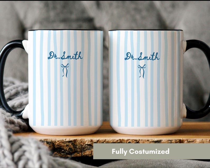 May include: Two white ceramic mugs with a light blue vertical striped pattern. Each mug features a black handle and the text Dr. Smith in dark blue script, accompanied by a small dark blue bow icon centered below the name on the front surface.