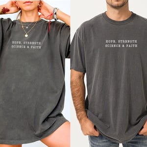 May include: A charcoal grey short-sleeve t-shirt featuring the white text HOPE, STRENGTH SCIENCE & FAITH printed on the chest. The garment has a crew neckline and a relaxed fit, showcasing a washed-out, vintage-inspired fabric texture.