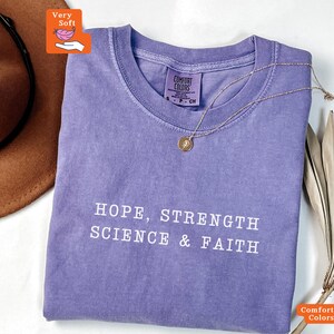 May include: A purple short-sleeved t-shirt with a crew neckline and the white text "HOPE, STRENGTH SCIENCE & FAITH" printed on the front. The shirt features a Comfort Colors brand label inside the collar and a thin gold-tone necklace.