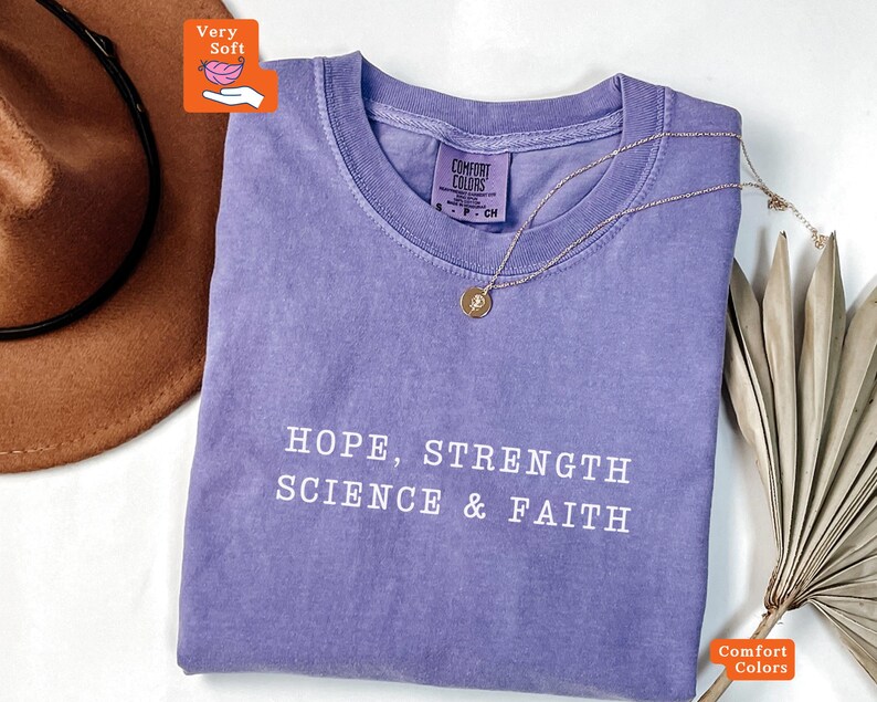 May include: A purple short-sleeved t-shirt with a crew neckline and the white text "HOPE, STRENGTH SCIENCE & FAITH" printed on the front. The shirt features a Comfort Colors brand label inside the collar and a thin gold-tone necklace.