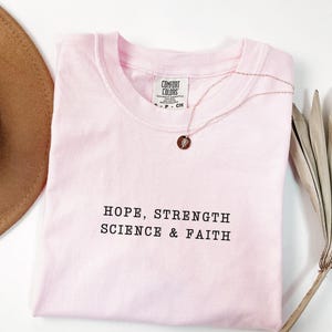 May include: A light pink short-sleeve t-shirt with a crew neckline featuring the black embroidered text HOPE, STRENGTH SCIENCE & FAITH. The shirt has a Comfort Colors brand label at the inner neck and is paired with a thin gold-tone necklace.