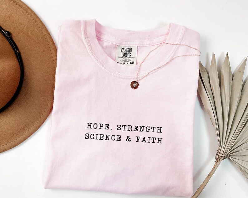 May include: A light pink short-sleeve t-shirt with a crew neckline featuring the black embroidered text HOPE, STRENGTH SCIENCE & FAITH. The shirt has a Comfort Colors brand label at the inner neck and is paired with a thin gold-tone necklace.