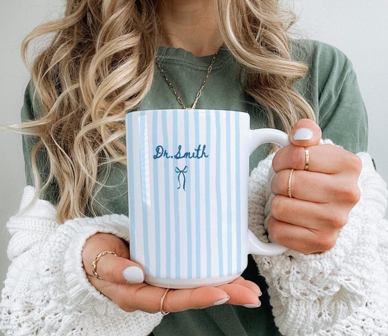May include: A white ceramic mug featuring vertical light blue stripes, the text Dr. Smith in blue script, and a small blue bow graphic. The mug has a curved handle and a smooth, glossy finish, providing a clean and simple design for daily beverage use.