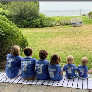 K&ouml;nnte beinhalten: Kinder tragen blaue Trikots mit wei&szlig;en Buchstaben und Zahlen und sitzen auf einer schwarz-wei&szlig; gestreiften Matte. Die Trikots zeigen Namen und Nummern. Der Hintergrund zeigt eine gr&uuml;ne Wiese, B&auml;ume und einen Blick auf das Meer.