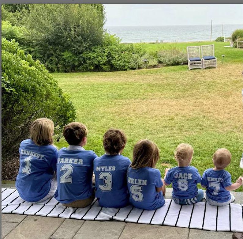 K&ouml;nnte beinhalten: Kinder tragen blaue Trikots mit wei&szlig;en Buchstaben und Zahlen und sitzen auf einer schwarz-wei&szlig; gestreiften Matte. Die Trikots zeigen Namen und Nummern. Der Hintergrund zeigt eine gr&uuml;ne Wiese, B&auml;ume und einen Blick auf das Meer.