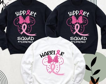 Puede incluir: Tres sudaderas con diseños de cinta rosa. Una sudadera blanca dice "Warrior" y las otras dos sudaderas negras dicen "Support Squad #TeamEmily".