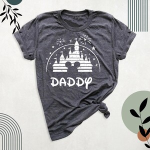 May include: Charcoal grey t-shirt with a white graphic depicting a castle, fireworks, and Mickey Mouse ears. The word "DADDY" is printed in a decorative font beneath the image. The shirt has a round neck and short sleeves.