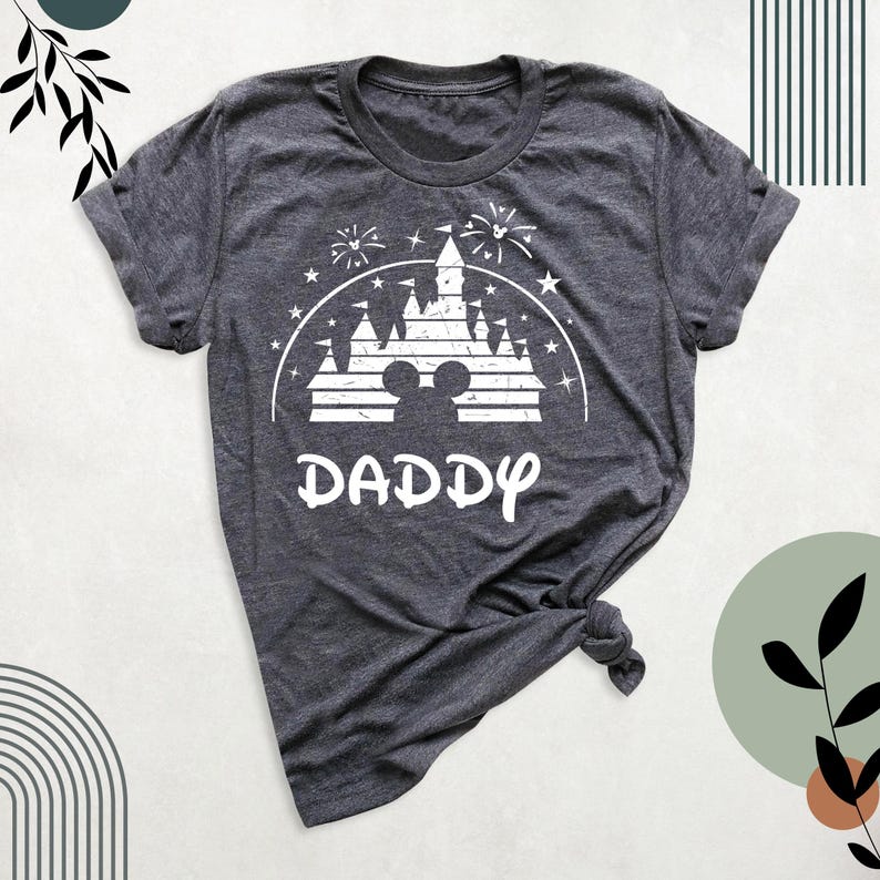 May include: Charcoal grey t-shirt with a white graphic depicting a castle, fireworks, and Mickey Mouse ears. The word "DADDY" is printed in a decorative font beneath the image. The shirt has a round neck and short sleeves.