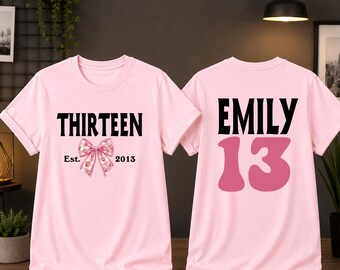 May include: A light pink t-shirt shown from the front and back. The front features the black text THIRTEEN, a pink strawberry-patterned bow, and the text Est. 2013. The back displays the name EMILY in black and the large pink number 13.