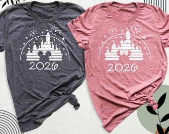 May include: A dark gray t-shirt and a dusty rose t-shirt, each featuring a white graphic of a castle silhouette, fireworks, stars, and the year 2026. The dark gray shirt includes a Mickey Mouse head icon, while the rose shirt features a Minnie Mouse head icon with a bow.