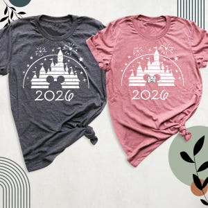 May include: Two short-sleeved t-shirts, one in charcoal grey and one in dusty rose. Each shirt displays a white graphic of a castle, fireworks, stars, and the year "2026". The grey shirt has Mickey Mouse ears, while the pink shirt has a bow.