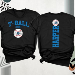 May include: Black T-shirts with baseball-themed designs. One shirt features "T-BALL MOM" and a baseball with the number 13. The other shirt displays "HARPER" vertically and a baseball with the number 14.