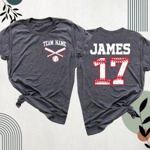 May include: Two gray t-shirts with a baseball theme. The front of one shirt has the text "TEAM NAME" with crossed baseball bats and a baseball. The back of the other shirt has the text "JAMES" and the number "17" in a baseball-stitched design.