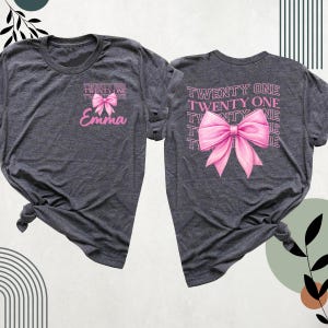 May include: Two grey t-shirts with the text "TWENTY ONE" and a pink bow. One shirt has the name "Emma" printed below the bow.
