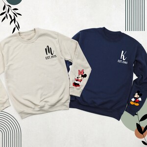 May include: Two long-sleeved sweatshirts, one beige and one navy blue. The beige sweatshirt has a black "M" and "EST. 1975" printed on the front. The navy blue sweatshirt has a black "K" and "EST. 1985" printed on the front. Both sweatshirts have a cartoon image of Mickey Mouse and Minnie Mouse on the sleeves.