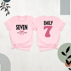 Peut inclure: Deux t-shirts roses, l'un avec le texte "SEVEN Est. 2018" et un n&oelig;ud rose, l'autre avec le texte "EMILY" et un grand chiffre 7 rose.