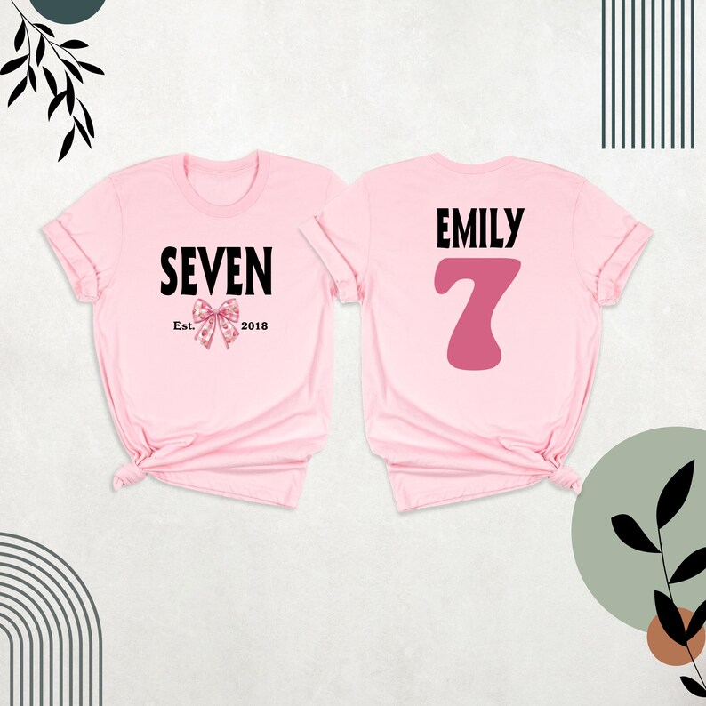 Peut inclure: Deux t-shirts roses, l'un avec le texte "SEVEN Est. 2018" et un n&oelig;ud rose, l'autre avec le texte "EMILY" et un grand chiffre 7 rose.