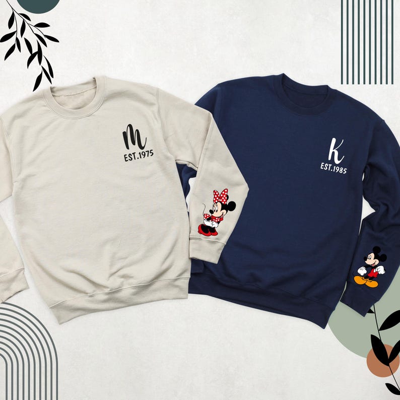 May include: Two long-sleeved sweatshirts, one beige and one navy blue. The beige sweatshirt has a black "M" and "EST. 1975" printed on the front. The navy blue sweatshirt has a black "K" and "EST. 1985" printed on the front. Both sweatshirts have a cartoon image of Mickey Mouse and Minnie Mouse on the sleeves.