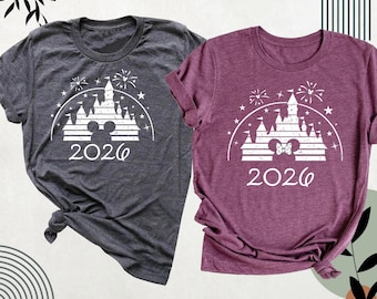 May include: A dark gray and a maroon short-sleeve t-shirt, each featuring a white graphic of a castle silhouette, fireworks, stars, and the year 2026. The gray shirt displays a Mickey Mouse head icon, while the maroon shirt shows a Minnie Mouse head icon with a bow.
