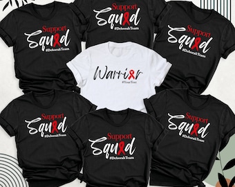 May include: Six black t-shirts with white text and a red ribbon graphic. The text on five of the shirts reads "Support Squad #DeborahTeam". The text on the sixth shirt reads "Warrior #YourText".