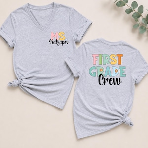 K&ouml;nnte beinhalten: Zwei graumelierte V-Ausschnitt-T-Shirts. Das linke T-Shirt zeigt den Text MS. Katzapov in Pink, Gelb und Schwarz. Das rechte T-Shirt zeigt den Text FIRST GRADE Crew in Pink, Orange, T&uuml;rkis und Schwarz mit kleinen orangefarbenen Blumen-Icons.