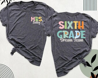 May include: A dark heather grey t-shirt shown from the front and back. The front features the text MRS. Bailey in pastel colours. The back displays the text SIXTH GRADE Dream Team in large, colourful block letters with small circular icons.