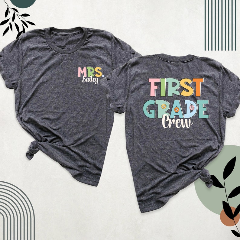 K&ouml;nnte beinhalten: Ein dunkelgrau meliertes T-Shirt, das von vorne und hinten gezeigt wird. Die Vorderseite tr&auml;gt den Schriftzug "MRS. Bailey" in Pink, Gelb und Wei&szlig;. Die R&uuml;ckseite zeigt den Text "FIRST GRADE Crew" in gro&szlig;en, bunten Blockbuchstaben mit kleinen Blumen.