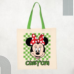 May include: A beige canvas tote bag with green fabric handles. The front features a graphic of Minnie Mouse wearing a red and white polka dot bow, centered over a green and white checkered square. The word CUSTOM is printed in green below.