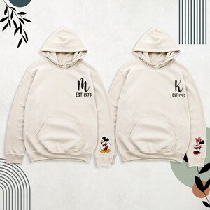 May include: Two cream-colored hoodies with Mickey Mouse and Minnie Mouse on the sleeves. The left hoodie has a black "M" with "EST. 1975" underneath. The right hoodie has a black "K" with "EST. 1985" underneath.