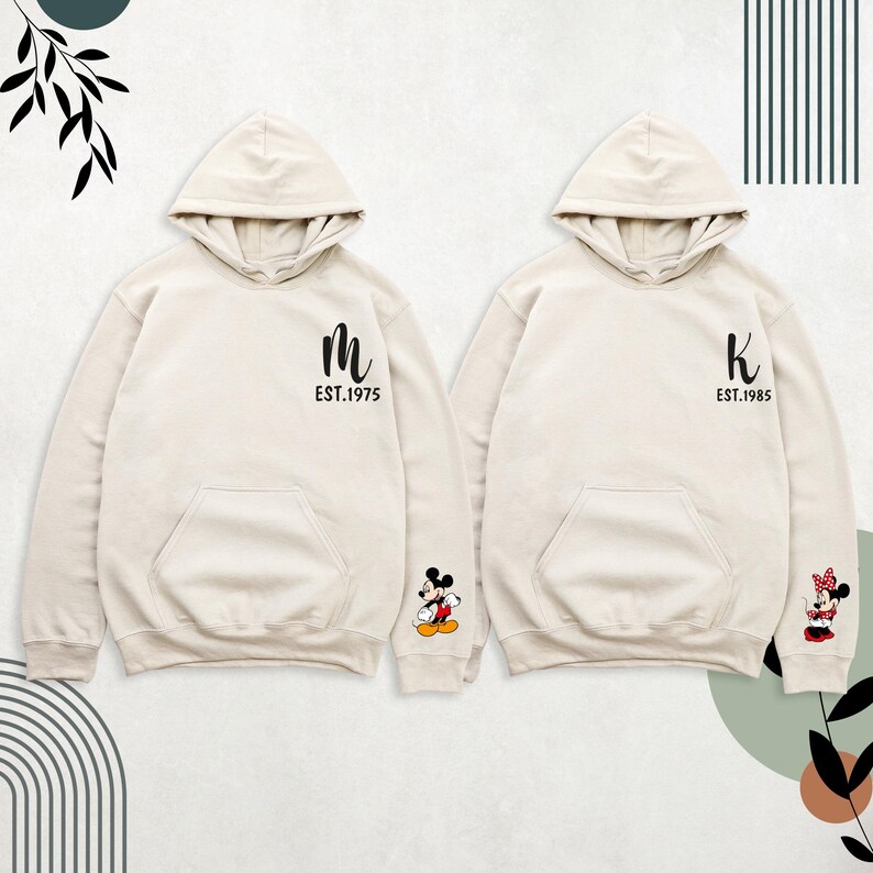 May include: Two cream-colored hoodies with Mickey Mouse and Minnie Mouse on the sleeves. The left hoodie has a black "M" with "EST. 1975" underneath. The right hoodie has a black "K" with "EST. 1985" underneath.