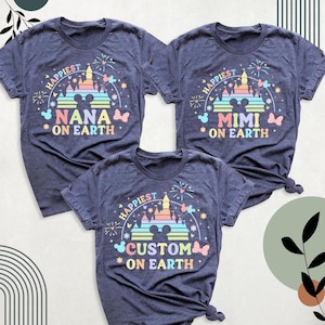 Puede incluir: Tres camisetas azul marino con un dise&ntilde;o gr&aacute;fico colorido. Cada camiseta presenta una silueta de castillo, fuegos artificiales y las palabras "Happiest" y "On Earth". Una camiseta dice "NANA", otra "MIMI" y la tercera "CUSTOM".